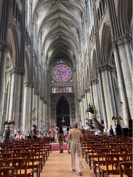 Interior of Reims cathedral, from the back of the church looking towards the altar. The cathedral is filling up with people. There is a colourful stained glass window high up in the back wall.