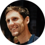 Chris Giles looking happy. He wears a plain dark blue shirt over a black t-shirt and has short brown hair and dark eyes.