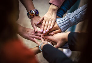 A close-up image of several hands of different skin tones coming together in the center of the frame. The hands are layered over each other. Visible on the wrists are various bracelets, adding a touch of individuality to each person.