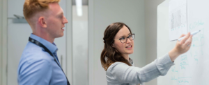 Female with long dark hair and glasses and wearing a grey top, writing on a white board. She is explaining to a man who is also looking at the board. He has short ginger hair and is wearing a plain blue shirt. He has a lanyard around his neck.