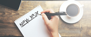 Woman writing New Year at the top of a blank page of a spiral bound notebook. On her desk is a cup of black coffee and a mobile phone.