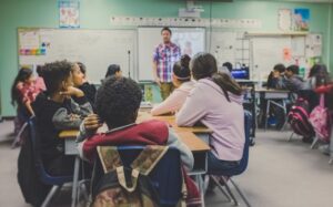 Male teacher stood at the front of the classroom with an interactive board with images of two children behind him. In the classroom are teenage children sitting around closets of tables.